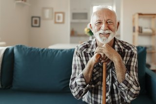 Älterer Mann lehnt sich auf dem Sofa sitzend auf seinen Gehstock und lächelt in die Kamera