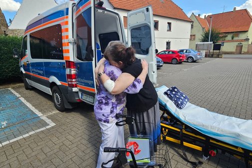 Tochter umarmt ihre Mutter herzlich, im Hintergrund steht ein Rettungswagen, eine Trage ist zu sehen.