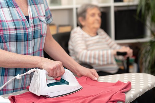 Seniorin im Rollstuhl, davor ein Bügelbrett und eine felissige Bügelhand im karierten Hemd.