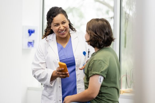 Ärztin lachend im Gespräch mit einer Patientin, diese hört gespannt zu, die Ärztin hält ein Smartphone in der Hand.