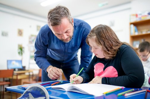eenager-M&auml;dchen mit Down-Syndrom in der Schule, sie zeichnet, ein Lehrer steht neben ihr und spricht mit ihr.