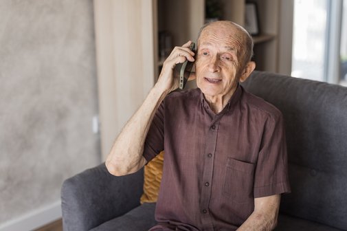 &Auml;lterer Mann sitzt auf dem Sofa und spricht in ein Telefon, das er mit der rechten Hand ans Ohr h&auml;lt.