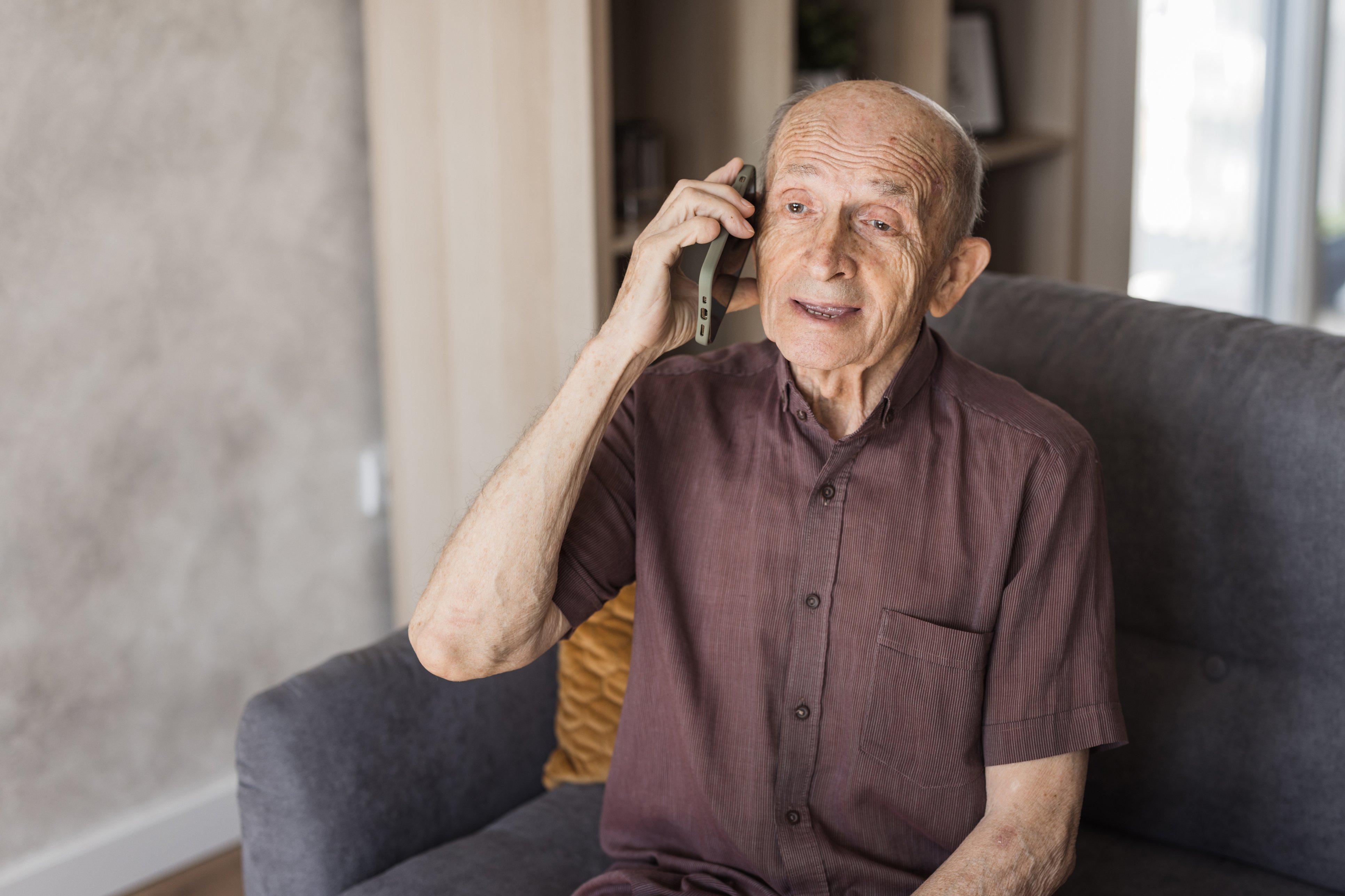 &Auml;lterer Mann sitzt auf dem Sofa und spricht in ein Telefon, das er mit der rechten Hand ans Ohr h&auml;lt.