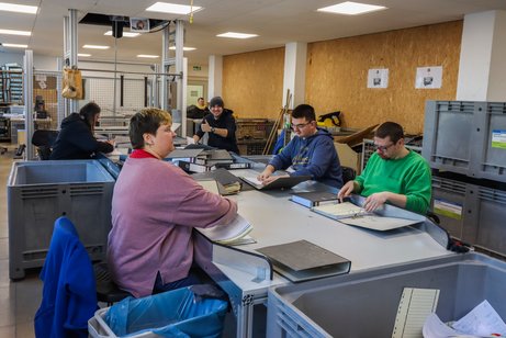 Aktenvernichtung Lebenshilfe Offenburg Arbeitssituation bei der Lebenshilfe, mehrere Mitarbeiter sitzen um einen Schreibtisch, vor sich zahlreiche Ordner, teilweise aufgeschlagen. Die Mitarbeiter blättern die Ordner durch und entfernen alles, was nicht Papier ist.