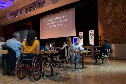 Blick auf die Zuschauer der SBV-Konferenz,eine Frau im Rollstuhl ist zu sehen, die einen Votrag verfolgt. Im Hintergrund die B&uuml;hne mit Leinwand.