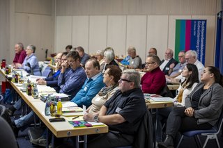 Seitenansicht auf eine Reihenbestuhlung, Menschen sitzen an ihren Plätzen und blicken nach vorne.