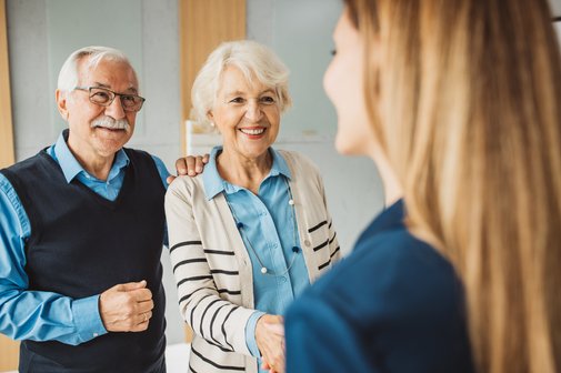 Begutachtung medizinischer Dienst Interview Ein älteres Paar steht lachend nebeneinander und begrüßt eine junge Frau, deren Gesicht noch rechts im Bild zu sehen ist.