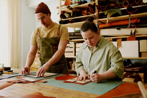 Arbeiten mit Menschen mit Behinderung Arbeit in einem Atelier, ein Mitarbeiter arbeitet mit seiner Kollegin mit Downsyndrom