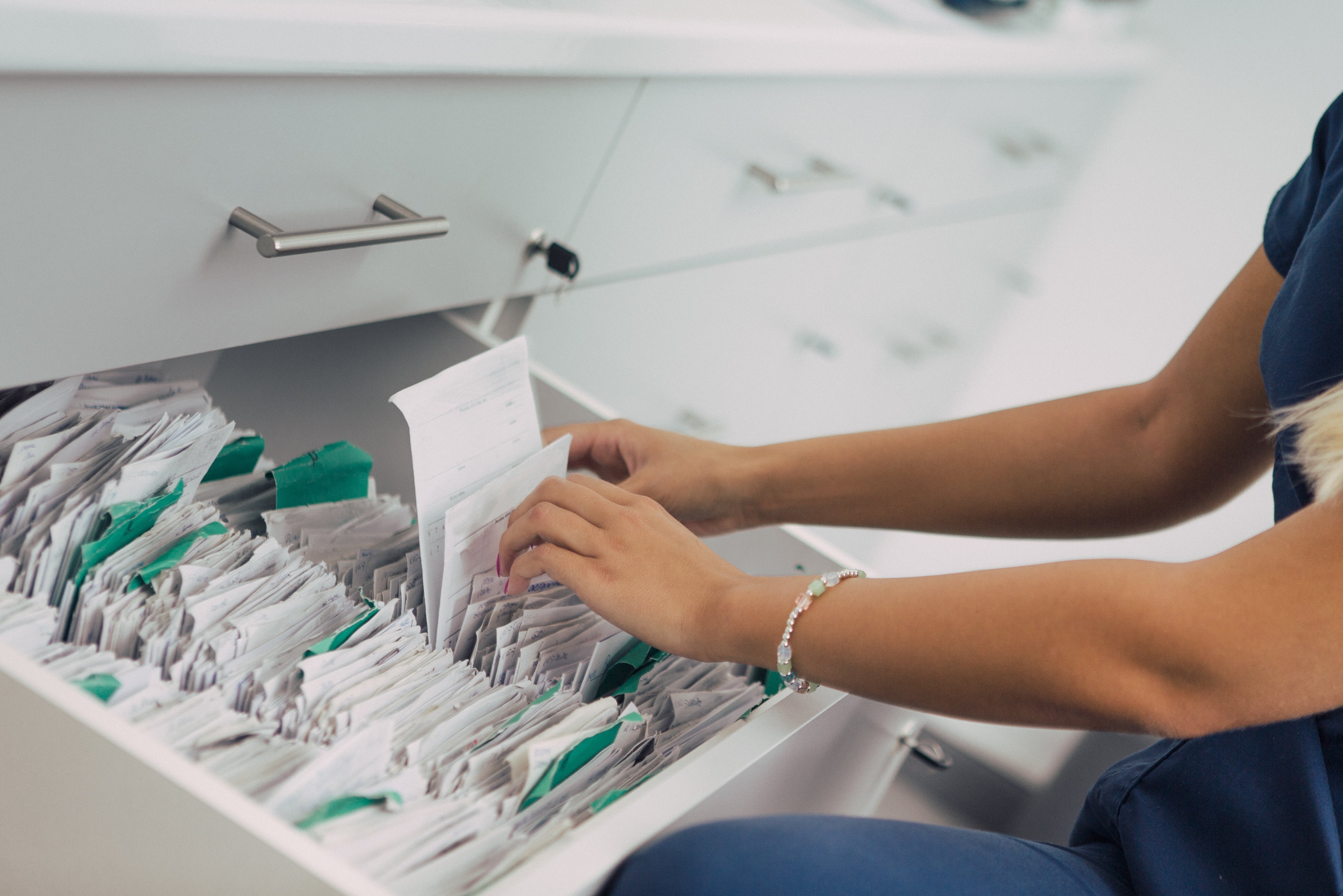 Geöffnete Schublade in einer Klinik, darin befinden sich mehrere Patientenakten, eine Krankenschwester sucht eine Akte heraus