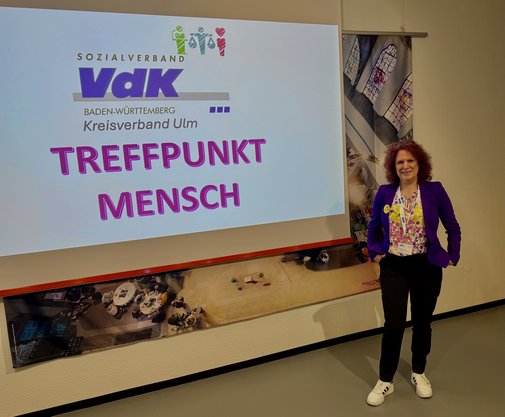 Renate Babic steht neben einer Leinwand, auf der "Treffpunkt Mensch" projiziert ist.