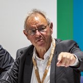 Drei M&auml;nner sitzen auf ihren Pl&auml;tzen am Podium und blicken auf die Delegierten vor ihnen.
