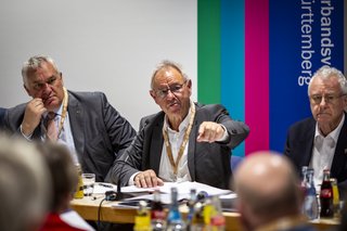 Drei Männer sitzen auf ihren Plätzen am Podium und blicken auf die Delegierten vor ihnen.