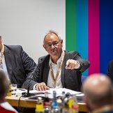 Drei M&auml;nner sitzen auf ihren Pl&auml;tzen am Podium und blicken auf die Delegierten vor ihnen.