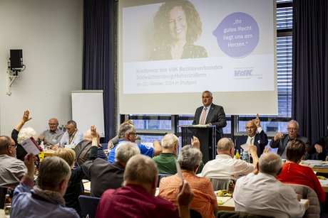 Innenansicht eines Saals, im Hintegrund auf der Leinwand ist eine Präsentation zu sehen, davor Jürgen Neumeister am Mikrofon, in der Menge ein paar Wortmeldungen.