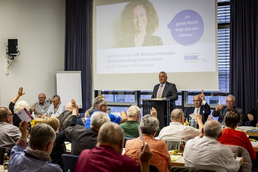 Innenansicht eines Saals, im Hintegrund auf der Leinwand ist eine Präsentation zu sehen, davor Jürgen Neumeister am Mikrofon, in der Menge ein paar Wortmeldungen.