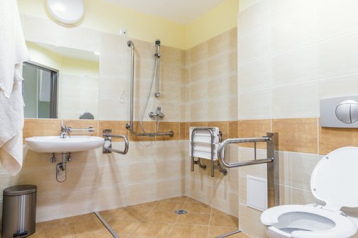Beispiel für ein barrierefreies Bad, die Dusche ist ohne Schwelle erreichbar, direkt nebe der Toilette ist ein Haltegriff, in der Dusche gibt es eine Sitzmöglichkeit. Das Waschbecken ist unterfahrbar.