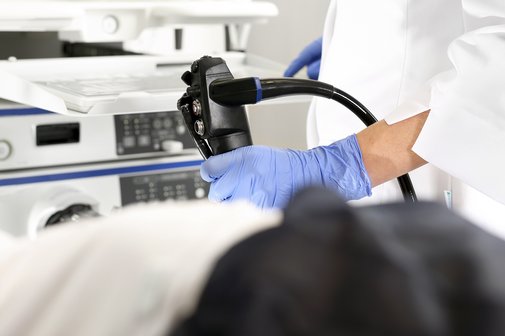 Darmspiegelung Vorsorge Untersuchung Darstellung einer Darmspiegelung mit einem Untersuchungsgerät, das von einem Arzt in den Händen gehalten wird, von diesem ist nur der Unterkörper und ein weißer Kittel zu sehen, im Vordergrund die Schemen eines liegenden Patienten.