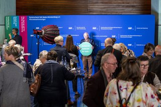 Pressewand im Hintergrund, davor eine Kamera und Licht aufgebaut, ein Mann hält eine Sprechblase mit der Aufschrift „Solidarität bedeutet für mich…“, im Vordergrund stehen viele Menschen und unterhalten sich.