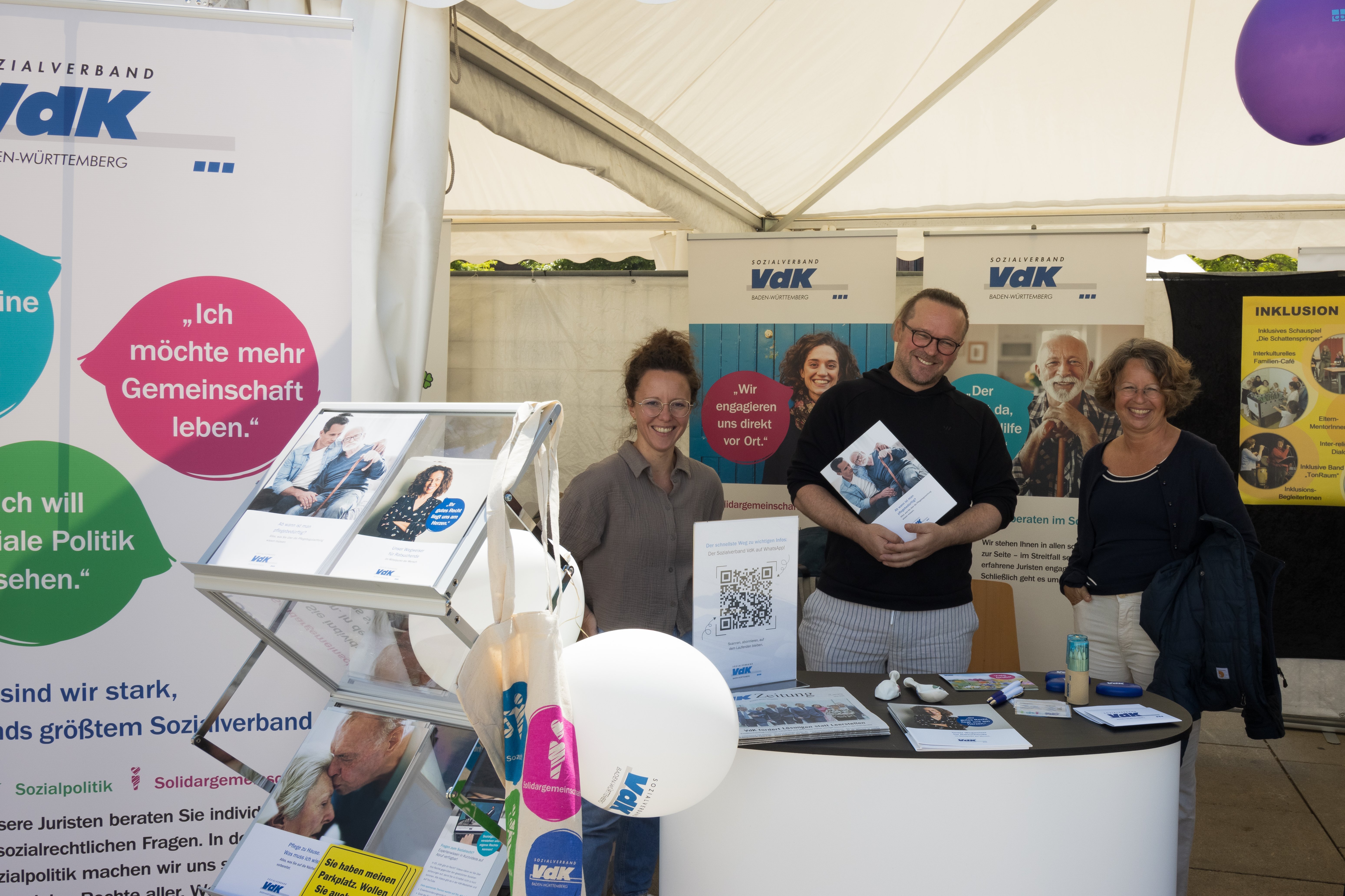 Aktionstag Inklusion 2025 Freiburg Blick von vorne auf den VdK-Infostand beim Aktionstag Inklusion in Freiburg, hinter einer kleinen Theke stehen drei Mitarbeiter und lachen in die Kamera. ThomasKupczyk steht in der Mitte und hält eine VdK-broschüre in der Hand.