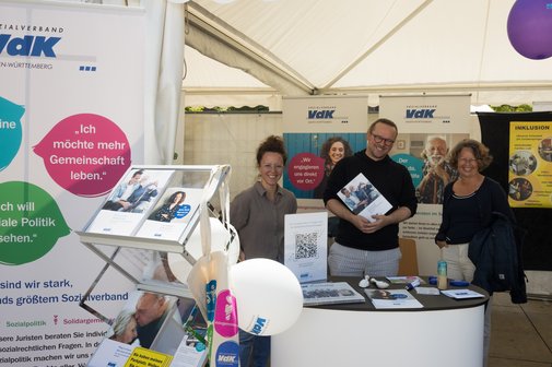 Aktionstag Inklusion 2025 Freiburg Blick von vorne auf den VdK-Infostand beim Aktionstag Inklusion in Freiburg, hinter einer kleinen Theke stehen drei Mitarbeiter und lachen in die Kamera. ThomasKupczyk steht in der Mitte und hält eine VdK-broschüre in der Hand.