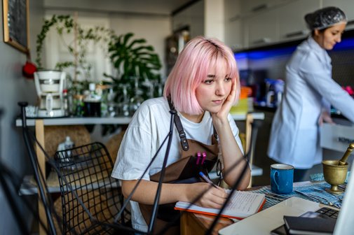 Studentin arbeitet in einem Caf&eacute; als Minijob, sie steht an der Theke und blickt, auf ihren Ellbogen gesch&uuml;tzt, auf die Kasse, rechts von ihr im Hintergrund steht eine Kollegin.