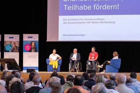 Blick auf die Bühne vom Publikum aus, vorne sind die Teilnehmer der Konferenz zu sehen, teilweise die Köpfe und Schultern. Auf der Bühne vier Diskussionsteilnehmer sitzend auf Stühlen im Gespräch miteinander. 