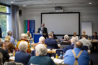 Innenansicht eines Saals, links Fenster. Die Rücken und Köpfen von verschiedenen Menschen an ihren Plätzen ist zu sehen, sie blicken auf Ernst Schilling am Pult, der eine Rede hält.