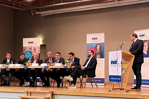 Vortrag und Podiumsdiskussion zur Landtagswahl Grünsfeld Blick auf eine Bühne, dort sitzen an einem Tisch mehrere Personen, jeweils mit einem Mikrofon vor sich. Rechts im Bild ein Rednerpult an dem ein weiterer Mann steht und spricht.