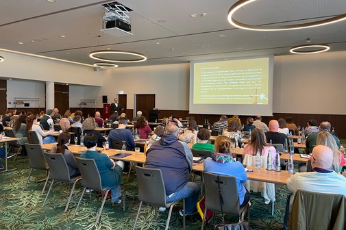 Schulungsraum innen, Lampen an der Decke, in Reihenbestuhlung mit Blick auf eine Leinwand, an der eine Pr&auml;sentation &uuml;bertragen wird, sitzen die Teilnehmer der Schulung auf ihren Pl&auml;tzen und verfolgen die Schulung.