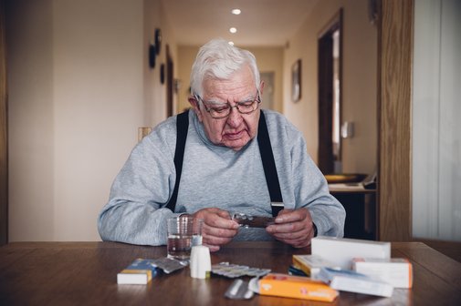 Medikamente Senior Rentner Management Älterer Mann sitzt am Tisch, er trägt eine Latzhose. Vor ihm liegen auf einem Tisch zahlreiche Tabletten, Tablettenverpackungen und leere Tablettendosen. Er liest angestrengt eine Packungsbeilage.