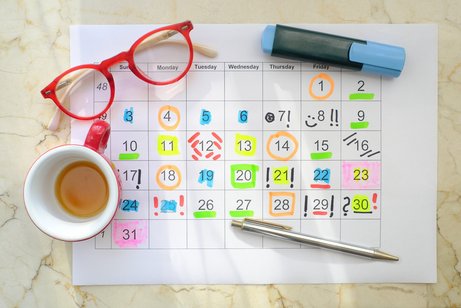 Monatskalender auf einem Papier, darauf liegen Stifte, daneben steht eine Kaffeetasse und eine Brille. Die Monatstage sind bunt markiert, eingekringelt und hervorgehoben mit verschieden farbigen Stiften.