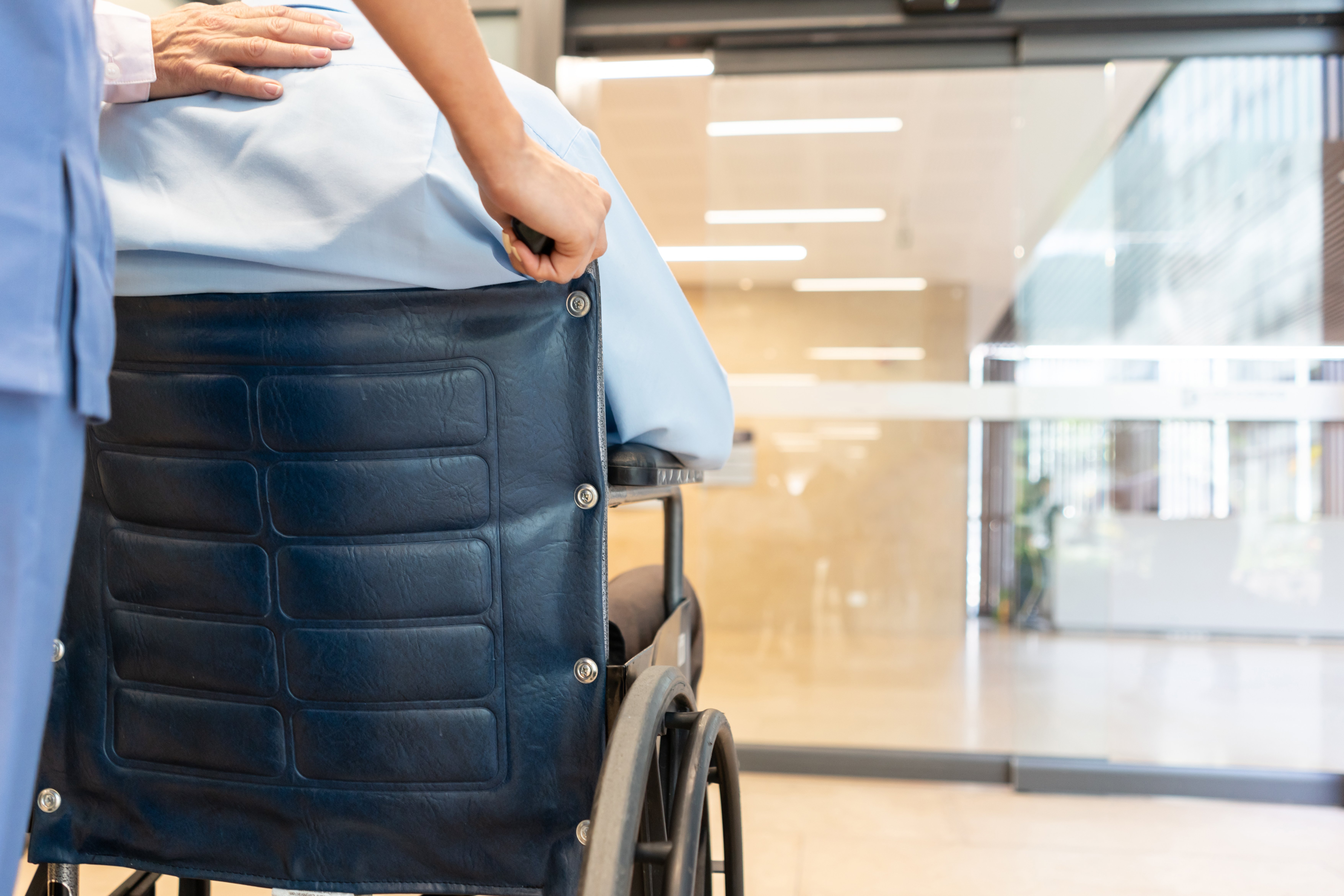 Nahaufnahme des nicht erkennbaren Mannes auf dem Rollstuhl im Krankenhaus und Krankenschwester schieben Rollstuhl, während Partner lehnt sich Hand auf Schulter