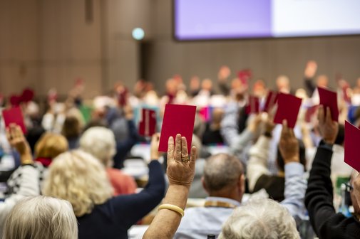 Innenaufnahme der Liederhalle in Stuttgart, Veranstaltungssaal, Nhaufnahme von hinten auf die Delegierten des Sozialverbandes VdK, ausgestreckte Hände sind zu sehen, die rote Stimmkärtchen halten, die Delegierten reihen sich auf ihren Plätzen aneinander, der Saal ist voll. 