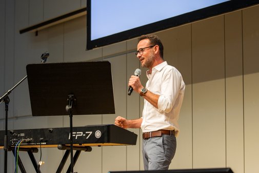 Jimmy Röck, Chorleiter und Dirigent, VdK-Gesundheitstag 2025 Jimmy Röck steht vor einem Piano auf der Bühne, mit einer Hand hält er ein Mikrophon, in das er gerade spricht