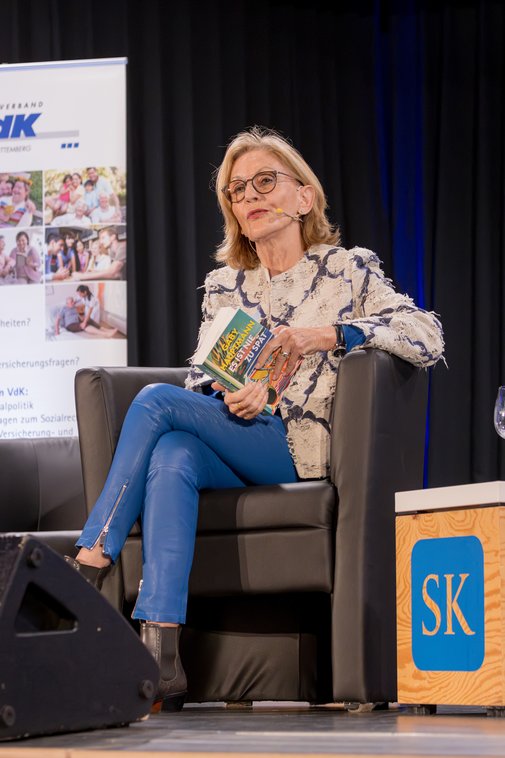 Gaby Hauptmann mit Brille sitzt in blauer Lederhose auf der B&uuml;hne und h&auml;lt ihr Buch &bdquo;Es ist nie zu sp&auml;t&ldquo; in den H&auml;nden, w&auml;hrend sie spricht. Neben ihr steht ein Beistelltisch mit dem Logo &bdquo;SK&ldquo;.