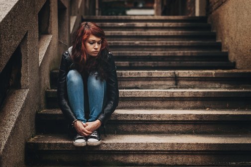 Junges Mädchen sitzt auf einer Treppe, sie umschlingt ihre Beine mit ihren Armen und blickt traurig auf die leeren Stufen neben sich.