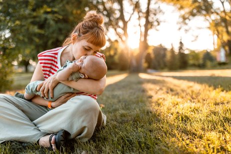 Junge Mutter sitzt mit ihrem Baby auf dem Rasen in der Natur, hält ihn im Arm und küsst ihn. Im Hintergrund sind Bäume und Sonnenschein zu sehen.