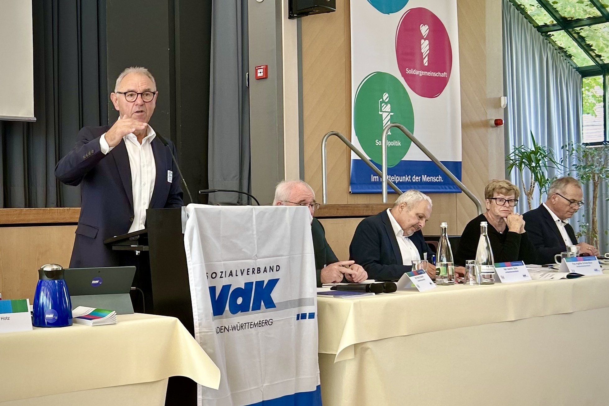 Ein Podium bei der Landesverbandskonferenz des Sozialverbands VdK Baden-W&uuml;rttemberg. Im Vordergrund h&auml;ngt eine wei&szlig;e Fahne mit dem blauen Logo des Verbands und dem Slogan &bdquo;Im Mittelpunkt der Mensch&ldquo;. Links spricht Hans-Josef Hotz im dunklen Sakko mit Brille an einem Rednerpult. Rechts daneben sitzen vier weitere Personen an einem langgezogenen, hell gedeckten Tisch und folgen der Veranstaltung.