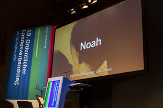 Ansicht einer Bühne mit Podium im Vordergrund, auf der Leinwand läuft ein Film mit Untertiteln.