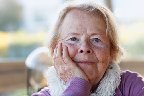Portrait von einer älteren Frau, sie lehnt sich auf ihren Arm und blickt in die Kamera.