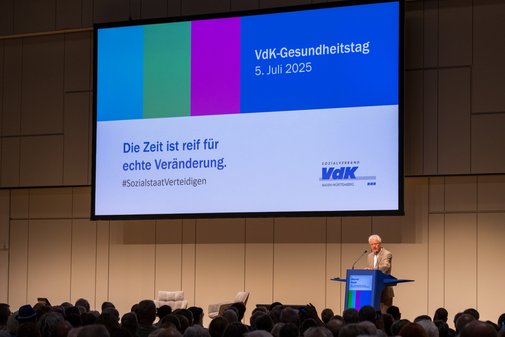 VdK-Gesundheitstag 2025 Werner Raab Blick auf die Bühne, Werner Raab steht am Rednerpult und spricht, im HIntergrund auf einer großen Leinwand ist die Aufschrift zu lesen"Die Zeit ist reif für echte Veränderung. #SozialstaatVerteidigen"