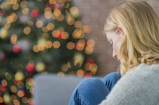 Allein zur Weihnachtszeit: Junge Fraue sitzt auf ihrem Sofa, die Beine angewinkelt und mit den Händen umklammert. Sie starrt traurig nach unten, im Hintergrund sind die Lichter eines Weihnachtsbaumes zu sehen.