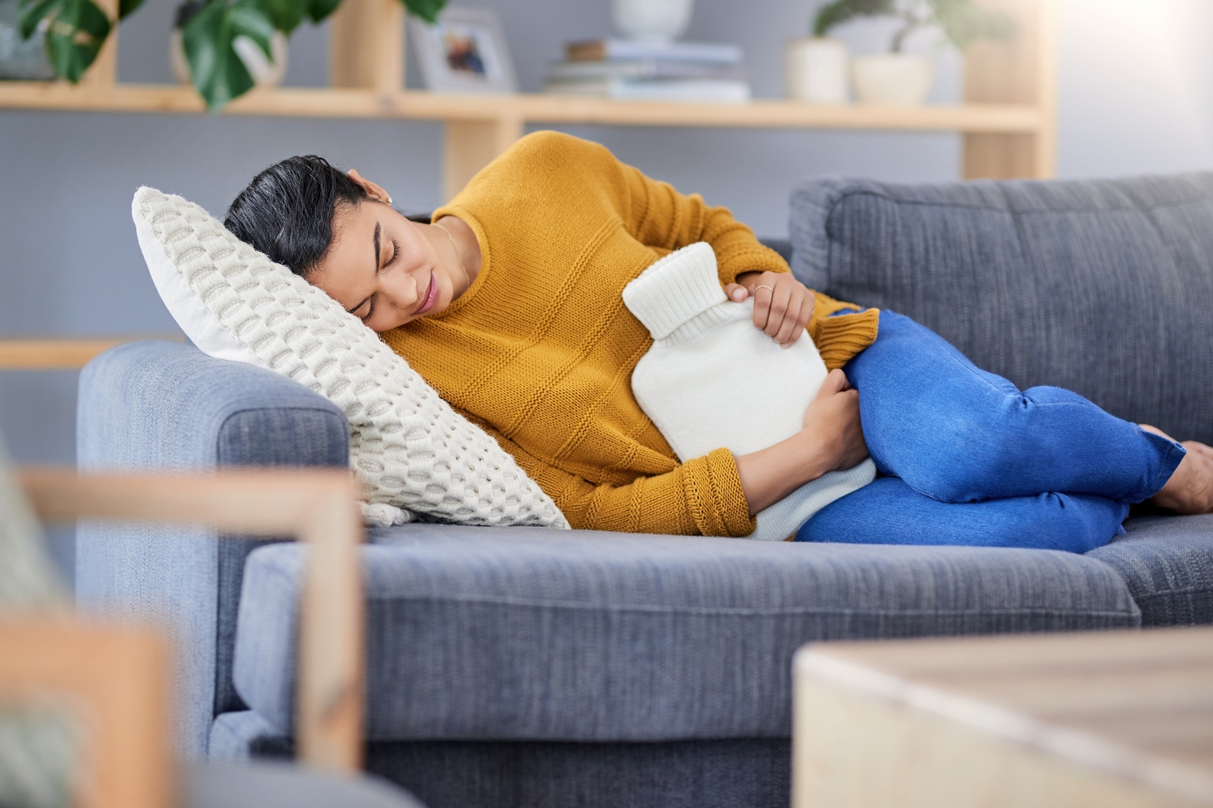 Frau mit schmerzverzerrtem Gesicht liegt seitlich auf dem Sofa, sie hält sich eine Wärmflasche an den Bauch.