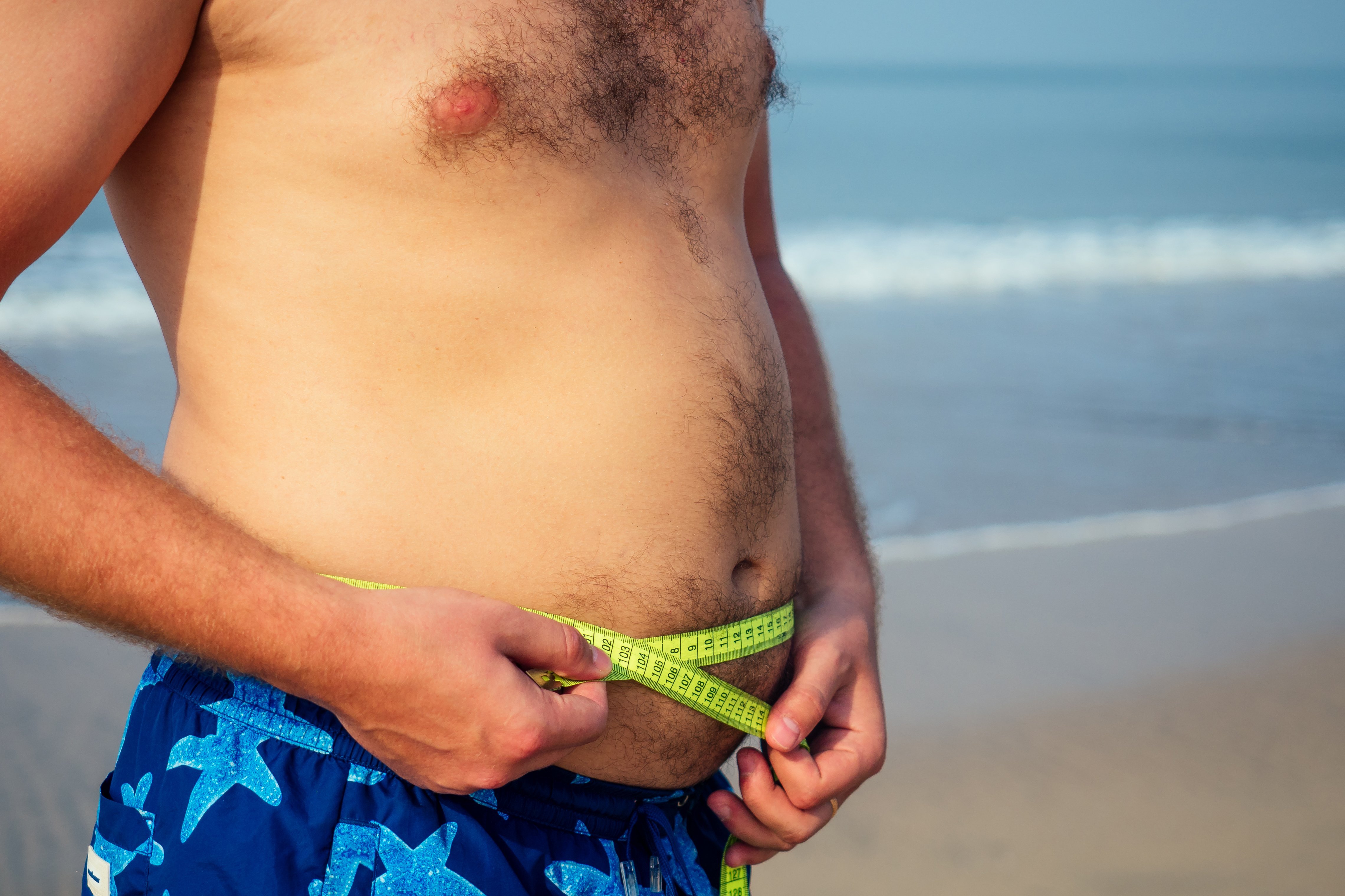 Mann in Badehose misst seinen Bauchumfang am Strand mit einem Schneidermassband