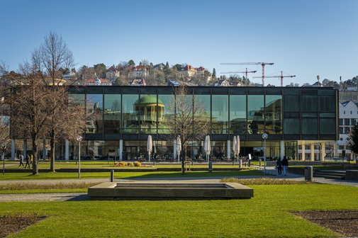Außenansicht des Landtags Baden-württemberg in Stuttgart: Ein gläsenernes Gebäude im Hintergrund, in dem sich ein gegenüberliegendes Gebäude mit einer Kuppel spiegelt. Vor dem Gebäude eine grüne Wiese und ein Brunnen. Im Hintergrund ist die Spitze eines Berges zu erkennen mit zahlreichen Häusern.
