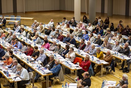 Reihenbestuhlung mit Stühlen und Tischen im Saal, die Delegierten sitzen auf ihren Plätzen und blicken auf die Bühne.