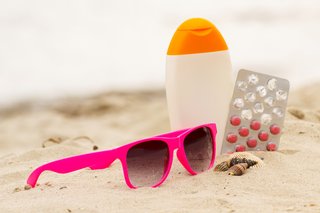 Rosafarbene Sonnenbrille liegt im Sand an einem Strand, daneben symbolisch eine Packung mit Medikamenten und eine Behälter mit Sonnencreme.