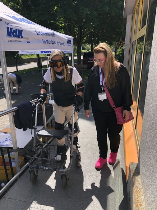 Links im Bild läuft Illona Jäger, sie trägt den Alterssimulationsanzug und versucht, mit einem Rollator zu gehen. Eine Mitarbeiterin am VdK-Stand unterstützt sie dabei.