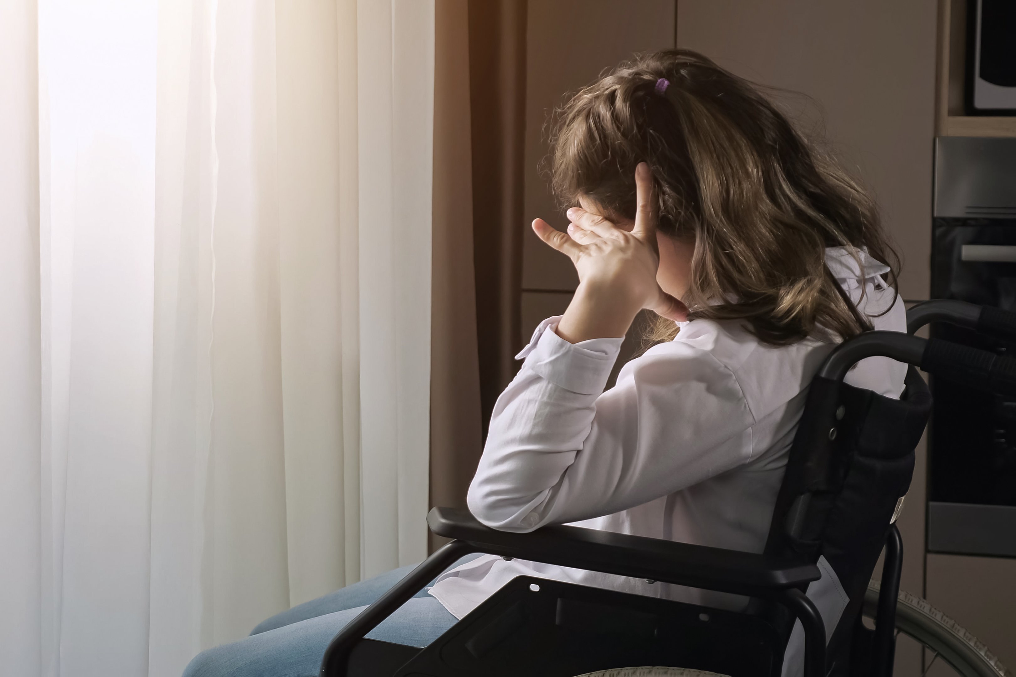 Eine junge Frau mit Behinderung sitzt in einem Rollstuhl an einem Fenster. Sie blickt nachdenklich oder sehns&uuml;chtig nach drau&szlig;en, w&auml;hrend warmes Sonnenlicht durch das Fenster f&auml;llt und sie sowie den Vorhang im Hintergrund beleuchtet.