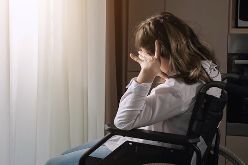 Eine junge Frau mit Behinderung sitzt in einem Rollstuhl an einem Fenster. Sie blickt nachdenklich oder sehns&uuml;chtig nach drau&szlig;en, w&auml;hrend warmes Sonnenlicht durch das Fenster f&auml;llt und sie sowie den Vorhang im Hintergrund beleuchtet.